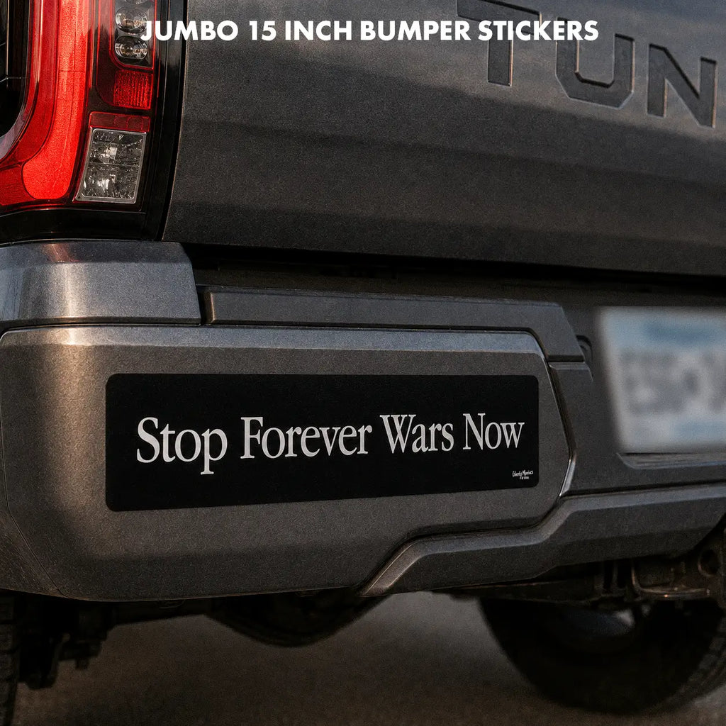 A black bumper sticker with the white text 'Stop Forever Wars Now' shown applied to the rear bumper of a dark gray pickup truck. White text at the top reads 'JUMBO 15 INCH BUMPER STICKERS'.