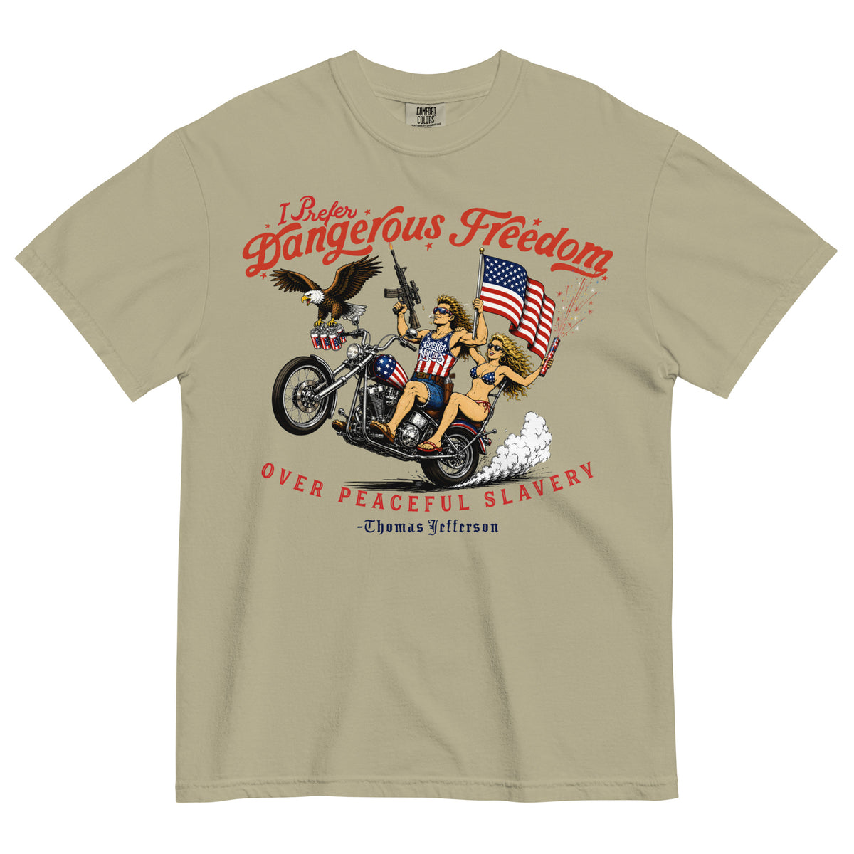 A beige short-sleeve t-shirt featuring a retro-style illustration of a man and women on a motorcycle with American flags, an eagle, and fireworks. Red text above reads 'I Prefer Dangerous Freedom' and text below reads 'OVER PEACEFUL SLAVERY -Thomas Jefferson'. By Liberty Maniacs