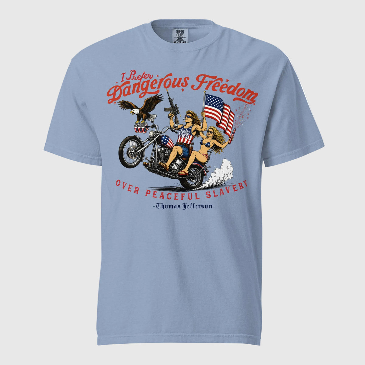 A light blue short-sleeve t-shirt featuring a retro-style illustration of a man and women on a motorcycle with American flags, an eagle, and fireworks. Red text above reads 'I Prefer Dangerous Freedom' and text below reads 'OVER PEACEFUL SLAVERY -Thomas Jefferson'. By Liberty Maniacs