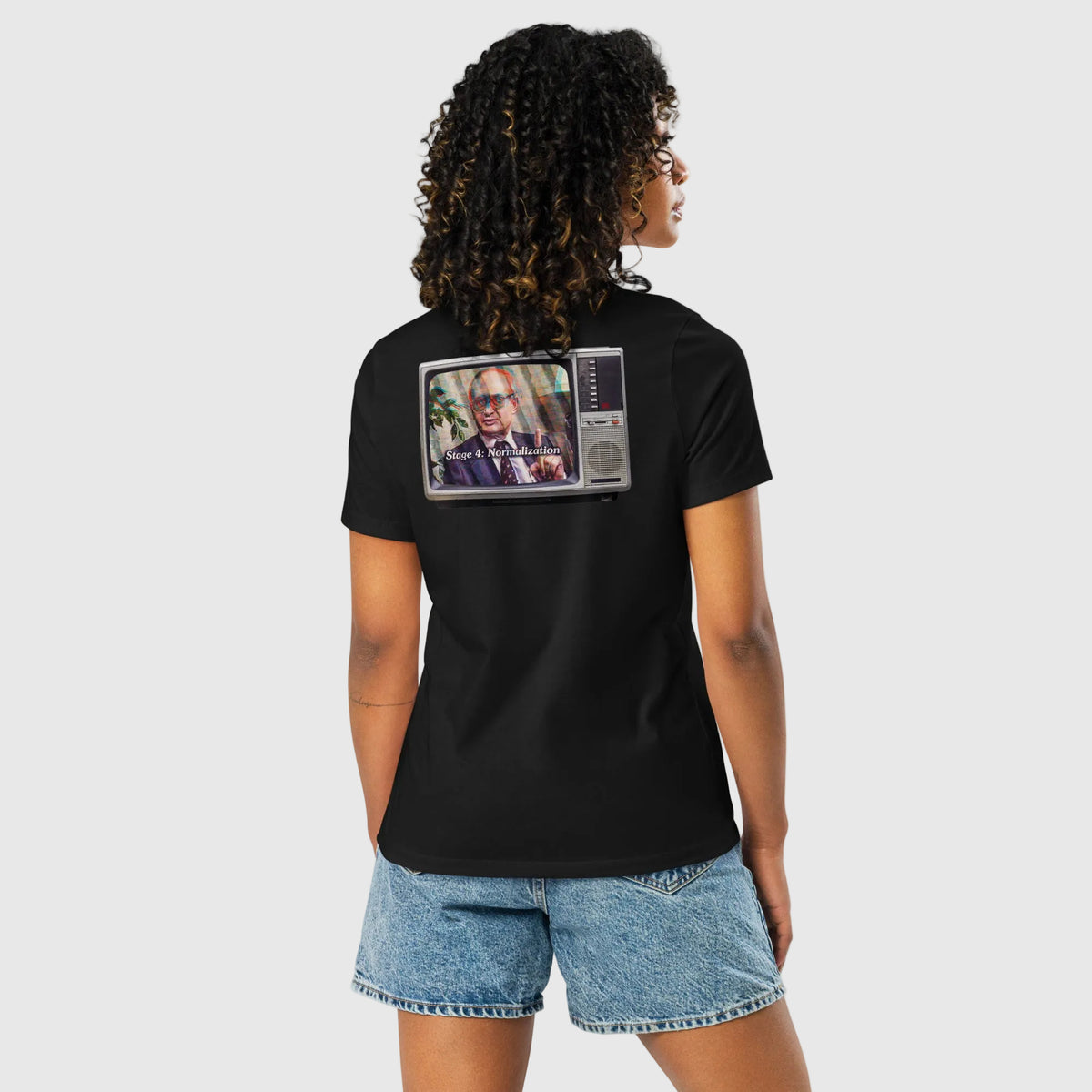A woman shown from the back wearing a black short-sleeve t-shirt with a graphic print of a vintage television showing a man with sunglasses. She is also wearing denim shorts against a plain white background.